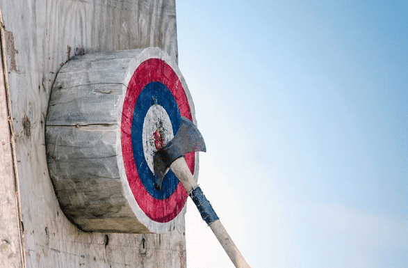 A vibrant and colorful half-cut tree with an axe stuck firmly into its trunk. A fun-filled atmosphere surrounds it, with people cheering and participating in an exciting axe throwing party. 